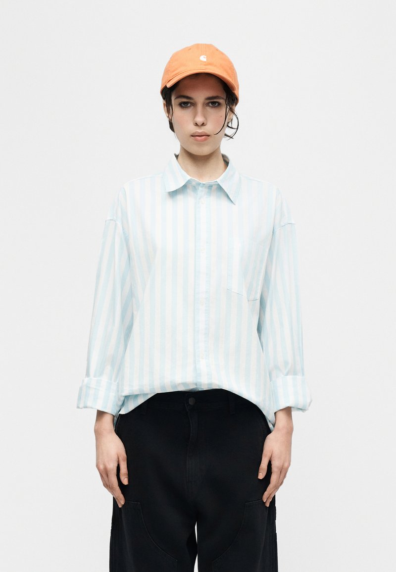 Persona che indossa un cappello arancione, una camicia a righe azzurre e bianche con bottoni e maniche arrotolate, e pantaloni neri larghi su uno sfondo semplice.