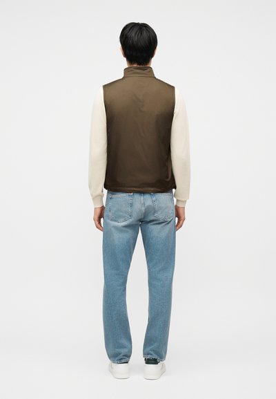 Άτομο που στέκεται με την πλάτη γυρισμένη φοράει καφέ αμάνικο γιλέκο πάνω από κρεμ πουκάμισο με μακριά μανίκια, μπλε τζιν και λευκά αθλητικά παπούτσια σε απλό φόντο.