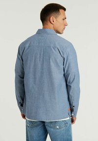 Camicia azzurra chiara con colletto, maniche lunghe, tessuto testurizzato, dettaglio nel yoke posteriore e cuciture a contrasto sui polsini. Abbinata a jeans sbiaditi.