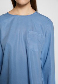 Femme portant un t-shirt uni bleu clair à manches longues avec un col rond et une petite poche poitrine sur le côté gauche.