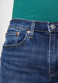 Primer plano de unos vaqueros de mezclilla azul con bolsillo frontal visible, trabilla para cinturón, remaches de cobre y botón, llevados con una blusa verde.