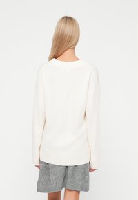 Krémový oversize sveter s rebrovaným výstrihom a manžetami, vyrobený z mäkkého materiálu, v kombinácii s textúrovanou sivou sukňou.