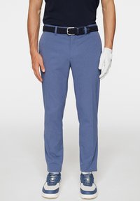 Hellblaue Hosen mit strukturiertem Gewebe, einem navyblauen gewebten Gürtel und weißen Handschuhen an einer Hand. Das Modell trägt marineblaue Schuhe mit blauen Akzenten.