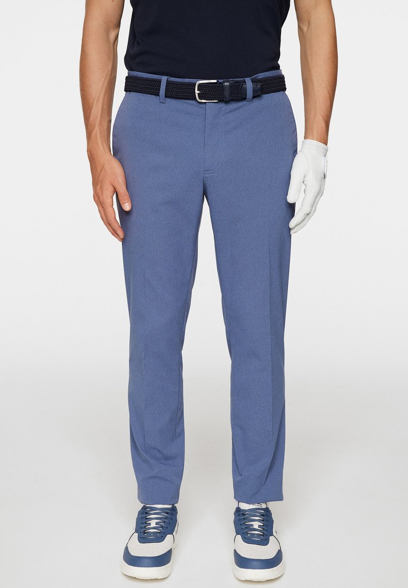 Hellblaue Hosen mit strukturiertem Gewebe, einem navyblauen gewebten Gürtel und weißen Handschuhen an einer Hand. Das Modell trägt marineblaue Schuhe mit blauen Akzenten.