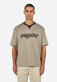 Beżowa koszulka z krótkim rękawem i dekoltem w serek, z ciemnobrązowym kołnierzykiem oraz słowem "pegador" napisanym grubą czarną czcionką na przodzie.