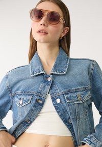 Svetlo modra denim jakna z strukturiranim ovratnikom, sprednjimi žepi in srebrnimi gumbi. Kombinirana z velikimi, toniranimi očali.