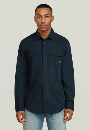 Mann mit kurzen Haaren, der ein dunkelblaues Hemd mit Brusttaschen und hellblaue Jeans trägt, steht vor einem einfarbigen hellen Hintergrund.