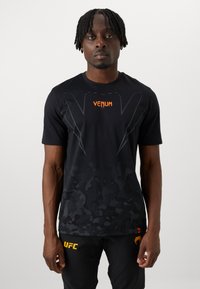 Camiseta deportiva negra con un patrón de camuflaje, logo "Venum" en naranja y detalles gráficos. Manga corta y diseño de cuello redondo.