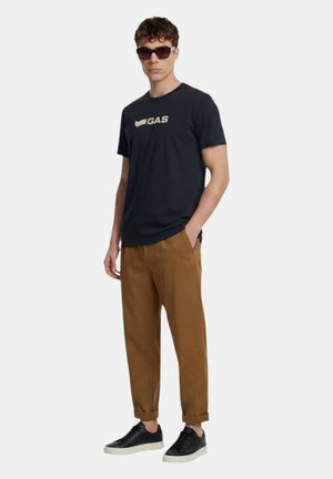Uomo che indossa occhiali da sole neri, t-shirt blu navy con logo, pantaloni marroni risvoltati e scarpe da ginnastica nere, in piedi con una mano in tasca.