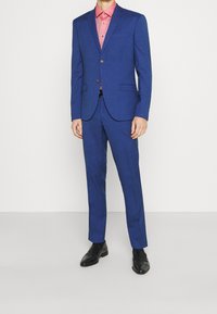Uomo che indossa un completo blu a due bottoni con pantaloni abbinati, una camicia a bottoni color salmone, una cintura nera, calzini neri e scarpe eleganti nere.