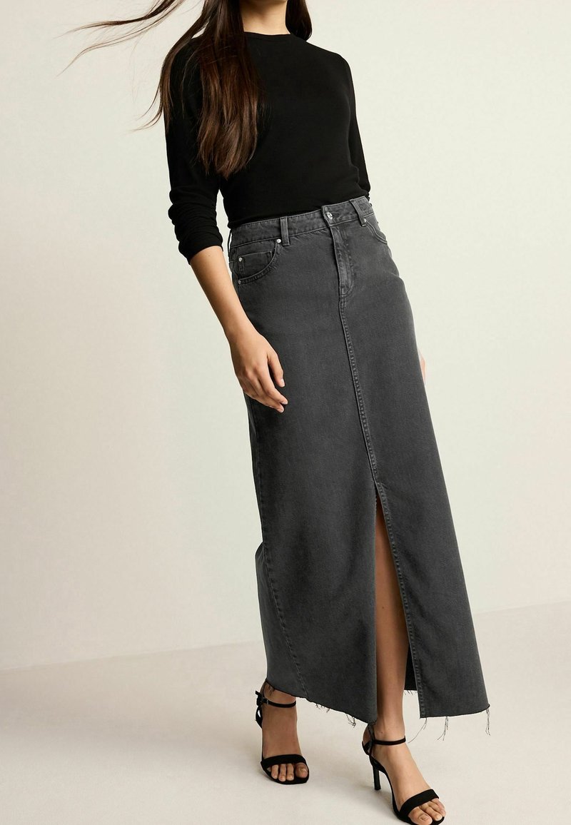 Falda larga de mezclilla gris de cintura alta con una abertura delantera, dobladillo sin rematar y cinco bolsillos. Combinada con una blusa negra de manga larga ajustada y tacones negros.