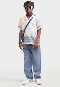 Krátky béžový rukáv s modrými geometrickými vzormi, modrý crossbody popruh, voľné svetlomodré jeansy a biele tenisky s hnedými akcentmi.