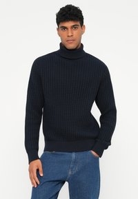 Pull en col roulé bleu marine en tricot texturé, avec des poignets et un ourlet côtelés, porté avec un jean bleu.