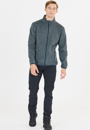 Graue zip-up Jacke mit hohem Kragen, ausgestattet mit zwei seitlichen Taschen und einem strukturierten Material. Kombiniert mit dunklen Hosen und schwarzen Stiefeln.
