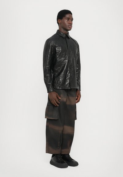 Black leather button-up shirt with a wrinkled texture paired with dark, wide cargo pants featuring a brown gradient and side pockets. Black sneakers.