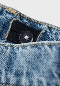 Tessuto denim con lavaggio blu, caratterizzato da una fascia elastica nera e un bottone di plastica trasparente con un motivo floreale. Dettagli di cucitura visibili.