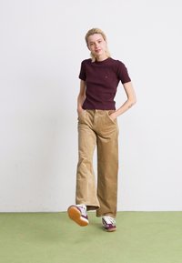 Giovane donna che indossa una camicia bordeaux, pantaloni in velluto a coste beige e scarpe da ginnastica bordeaux e bianche, in piedi con le mani nelle tasche e un piede sollevato.