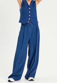 Gilet bleu avec cinq fermetures à boutons, assorti à un pantalon à jambes larges dans le même tissu et la même couleur. Texture lisse sans motifs visibles.