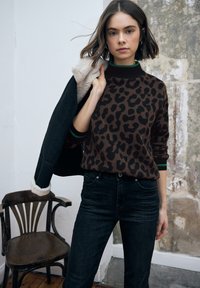 Brązowy sweter w nadruk lamparci z zielonymi akcentami, zestawiony z czarnymi dżinsami. Model trzyma czarną kurtkę z puszystym kołnierzem. W tle drewniane krzesło.