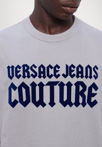 Tricou din bumbac gri cu un text bold de culoare albastru marin: "VERSACE JEANS COUTURE." Guler rotund și mâneci scurte cu o textură netedă.