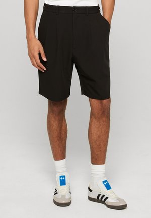 Homme portant un short noir sur mesure avec des plis, des chaussettes blanches et des baskets blanches avec des bandes noires et des écussons de logo bleus.