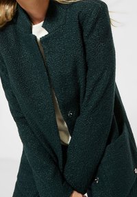 Cappotto verde scuro con trama, dal design senza colletto, dotato di chiusura a bottone e tasche laterali, realizzato in un morbido materiale peloso.