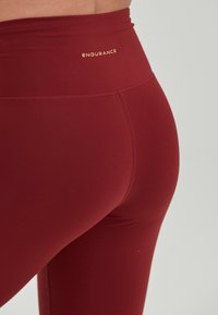 Hooggeschoren bordeauxrode leggings gemaakt van rekbare stof. Bevat een gouden "ENDURANCE" logo op de tailleband. Zachte textuur en aansluitend ontwerp.