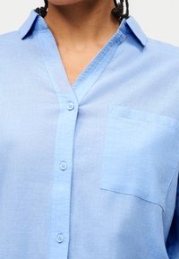 Άτομο που φοράει ανοιχτό μπλε πουκάμισο με γιακά, κουμπιά στο μπροστινό μέρος και τσέπη στο στήθος, με εμφανή το λαιμό και το ανώτερο μέρος του σώματος.