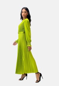 Lange, geplooide jurk in levendig limoengroen met een nauwsluitend lijfje en wijd uitlopende mouwen, voorzien van een ceintuur in de taille en een gestructureerde stof. Zwarte hakken.
