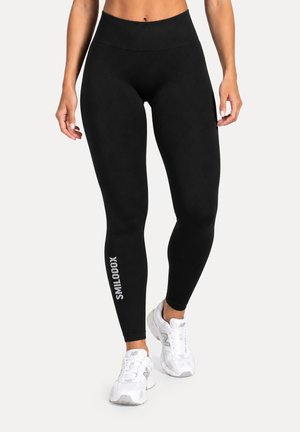 Femme portant des leggings noirs taille haute avec le texte "SMILODOX" sur la jambe gauche et des chaussures de sport blanches, debout devant un fond uni.