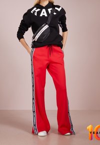 Černá mikina s kapucí s nápisem "KARL", doplněná jasně červenými tepláky s bočními pruhy s názvem značky v černé a bílé barvě. Oblečení doplňují bílé tenisky.