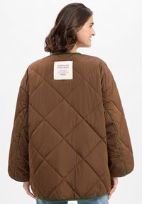 Smeđi quiltani kaput s zelenim obrubom, s pravokutnom oznakom na leđima. Dizajn uključuje dijamantni uzorak i široke rukave.