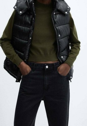 Person trägt eine schwarze Steppweste über einem olivgrünen kurzen Pullover mit den Händen in den Taschen schwarzer hoch taillierter Jeans.