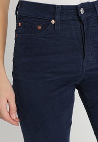 Gros plan sur une personne portant un pantalon en velours côtelé bleu foncé avec des poches avant et des rivets en cuivre, main détendue sur le côté.