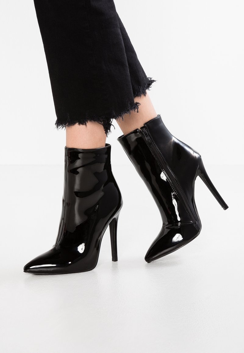 Bottines en cuir verni noir brillant avec bouts pointus, talons aiguilles hauts, fermetures éclair latérales, portées avec un pantalon noir raccourci à ourlets effilochés.