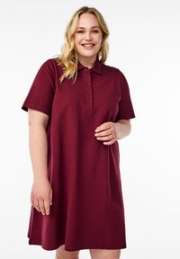 Bordeaux polo jurk met korte mouwen, een kraag en knoopsluiting. Gemaakt van zachte, textuurstof en heeft een ontspannen pasvorm en een lengte tot de knie.