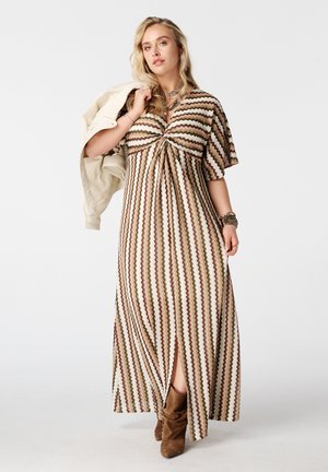 Vrouw draagt een lange bruine en beige jurk met een zigzagpatroon, houdt een lichte jas over de schouder en draagt bruine suède laarzen.