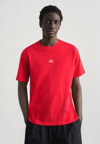 Νέος άνδρας με σκούρο δέρμα και κοντά, στριφτά μαλλιά, φοράει κόκκινο μπλουζάκι με μικρό λευκό λιοντάρι στο στήθος, με τα χέρια στις τσέπες μαύρων σορτς.