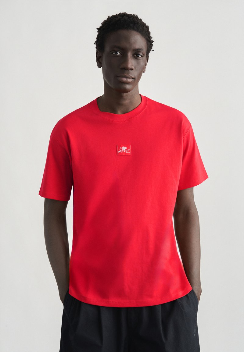 Νέος άνδρας με σκούρο δέρμα και κοντά, στριφτά μαλλιά, φοράει κόκκινο μπλουζάκι με μικρό λευκό λιοντάρι στο στήθος, με τα χέρια στις τσέπες μαύρων σορτς.