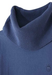 Pull à col montant bleu marine en tricot lisse, avec un col haut et une texture côtelée. Comprend un petit emblème sur le côté inférieur.