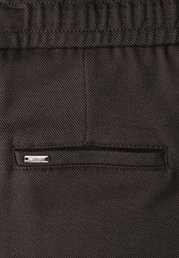 Brauner Strukturstoff mit einem gerippten Muster, versehen mit einer kleinen rechteckigen Tasche mit einem silbernen "Street One"-Logoschild.