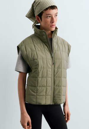 Personne portant une chemise à manches courtes gris clair, un gilet matelassé sans manches vert olive, un pantalon noir et un foulard clair rayé sur la tête, tourné vers la droite.