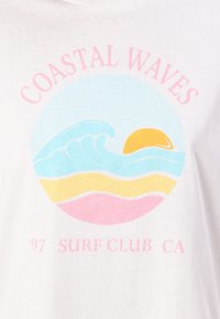 Balta T-kreklā ar apļveida grafiku, kurā ir zilas viļņi, dzeltena saule un slāņi pasteļu krāsās. Uzraksts: "COASTAL WAVES 97 SURF CLUB CA."