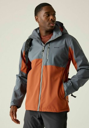 Man wearing a two-tone gray and orange outdoor jacket with hood and backpack, looking to his right with one hand in pocket.
