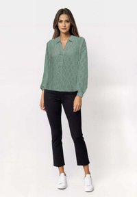 Camicia verde chiaro a fantasia con una vestibilità rilassata, chiusura a bottoni e maniche lunghe, abbinata a pantaloni neri e sneakers bianche.