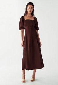 Rjava obleka srednje dolžine s kvadratnim izrezom, napihnjenimi rokavi in prilegajočim pasom. Gladka tekstura in minimalističen dizajn. Nosi se z bež platformskimi sandali.