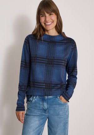 Femme souriante portant une chemise à carreaux bleue et noire manches longues et un jean bleu clair, debout, une main dans la poche, sur un fond uni.