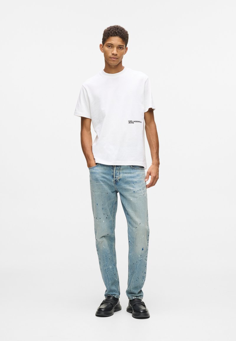 Λευκή βαμβακερή μπλούζα με ένα μικρό μαύρο λογότυπο, συνδυασμένη με ανοιχτό μπλε τζιν με σχέδια από splatter μπογιάς. Κλασικά μαύρα παπούτσια ολοκληρώνουν την εμφάνιση.