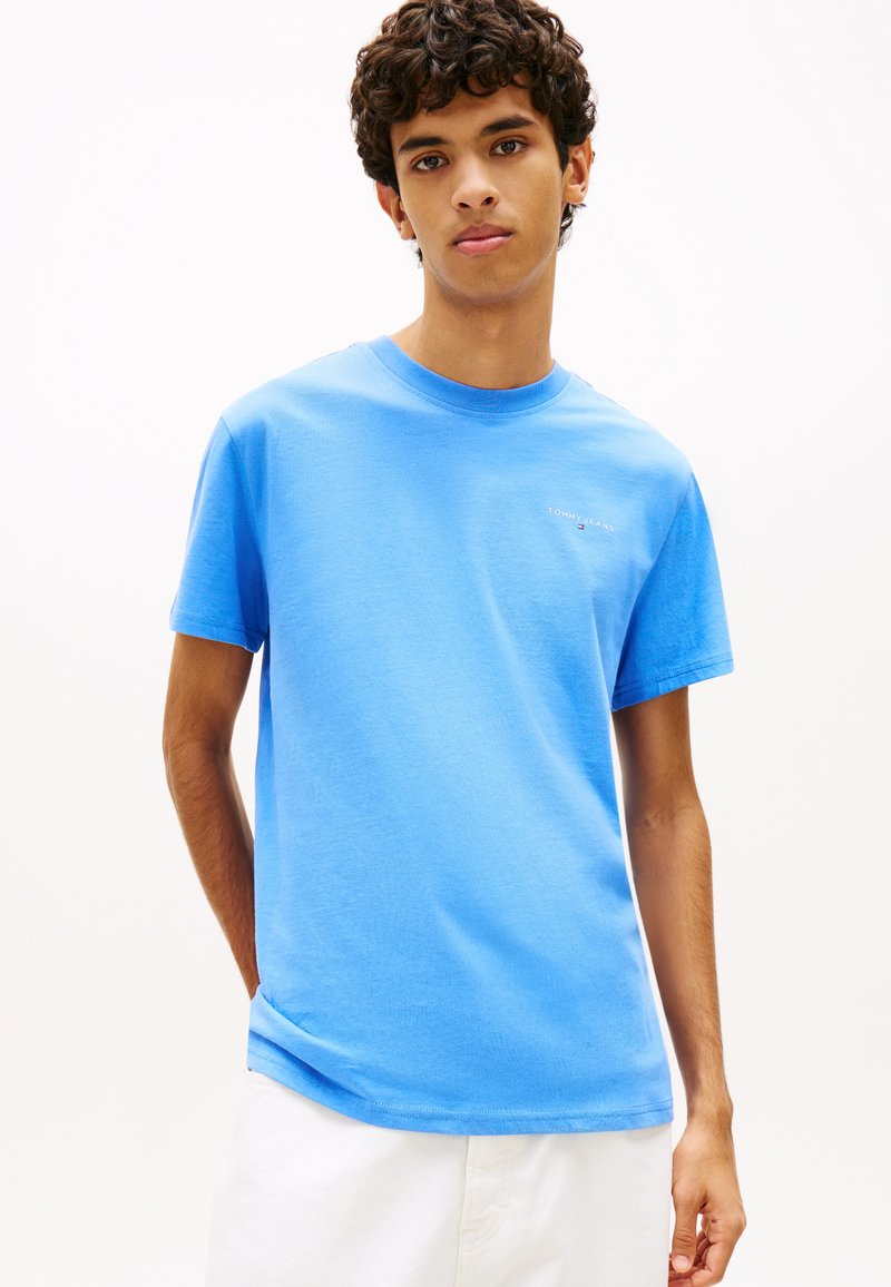 Νεαρός με κατσαρά μαλλιά φοράει ανοιχτό μπλε μπλουζάκι Tommy Jeans και λευκό παντελόνι σε λευκό φόντο.