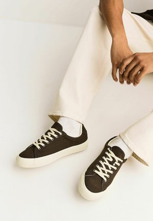 Asmuo dėvi smėlio spalvos kelnes ir juodus žemų batų sportbačius su baltais raišteliais ir pado dalimis, sėdi, rankas padėjęs ant kelių.